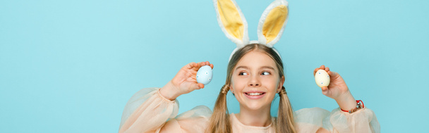 панорамный снимок веселого ребенка с кроличьими ушами, держащего окрашенные пасхальные яйца и смотрящего в сторону изолированного на голубом
  - Фото, изображение