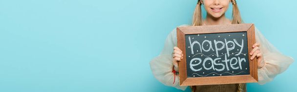 panoramic shot of cheerful kid holding chalkboard with happy easter lettering isolated on blue  - Fotó, kép