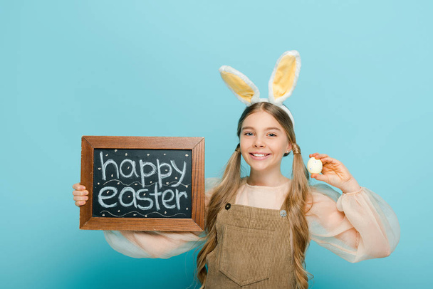 mosolygós gyerek nyuszifül gazdaság tábla boldog húsvéti betű és festett csirke tojás elszigetelt kék  - Fotó, kép