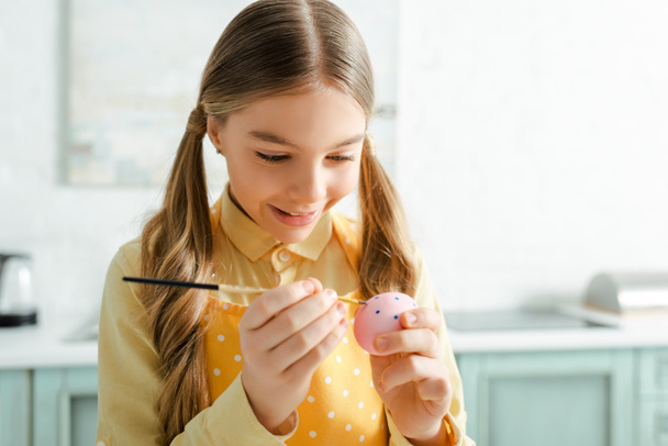 happy kid painting easter egg with paintbrush at home - Фото, зображення