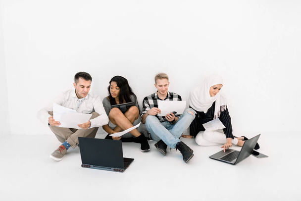 Group of Four young creative Multiethnic People Planning a New Project, talking each other, using laptop and tablet, while sitting on the floor on white background - Фото, изображение