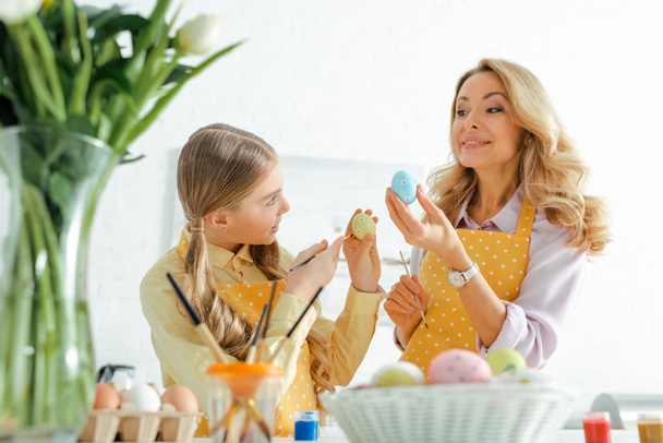 selective focus of happy mother and daughter holding paintbrushes and painted easter eggs  - Foto, imagen
