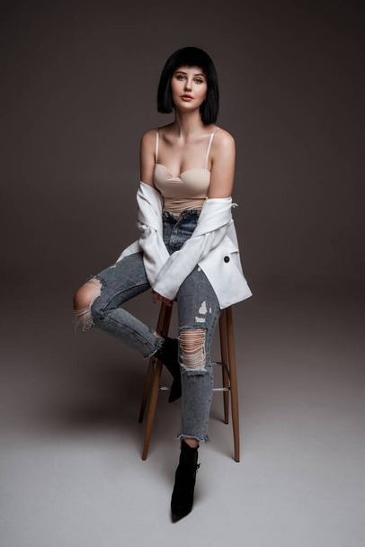 young beautiful brunette with short haircut. model wearing white jacket and jeans. lady in casual clothes posing on grey studio background. model on bar chair. fashion industry - 写真・画像