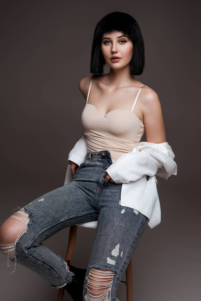 young beautiful brunette with short haircut. model wearing white jacket and jeans. lady in casual clothes posing on grey studio background. model on bar chair. fashion industry - Photo, Image