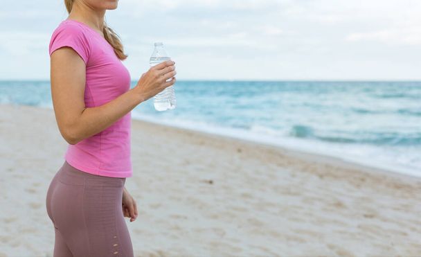 Attractive slim Woman drinking water in bottle after exercises on the beach in sports wear. Healthy lifestyle People Concept. Copy Space - Zdjęcie, obraz