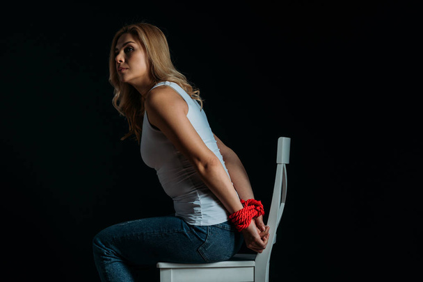 Vista laterale della donna con le mani legate che distolgono lo sguardo, seduta sulla sedia isolata sul nero
 - Foto, immagini