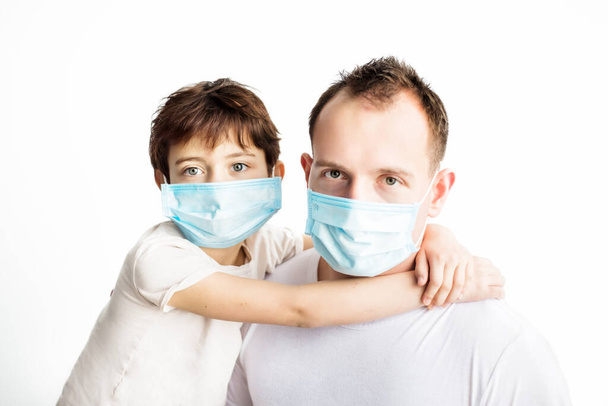Παιδί και μπαμπάς με μπλε ιατρικές μάσκες και λευκά μπλουζάκια αγκαλιάζονται και κοιτάζονται στην κάμερα σε λευκό φόντο. Προστασία από τον ιό του κερατοειδούς. Ιατρική περίθαλψη. Πρόληψη ασθενειών covid-2019 - Φωτογραφία, εικόνα