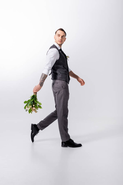 side view of handsome tattooed elegant bridegroom walking with bouquet on grey - Photo, Image