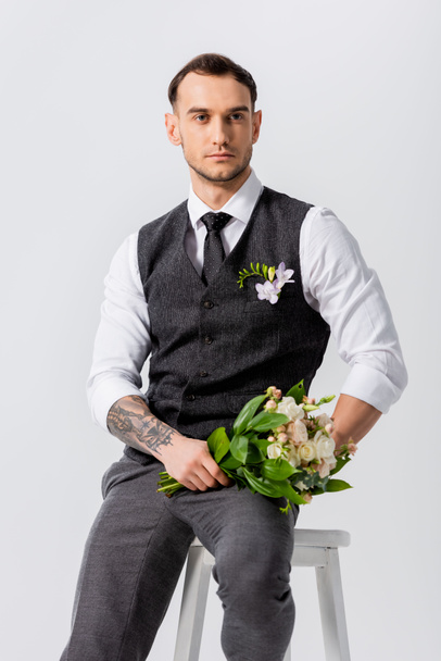 handsome tattooed elegant bridegroom with bouquet sitting on chair isolated on grey - Fotó, kép