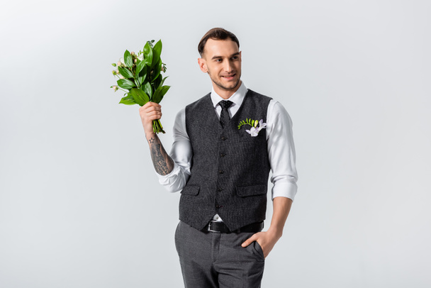 smiling handsome tattooed elegant bridegroom with bouquet and hand in pocket isolated on grey - Photo, Image