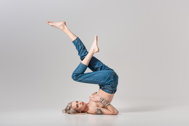 young topless tattooed girl in jeans posing on floor with raised legs on grey - Фото, изображение