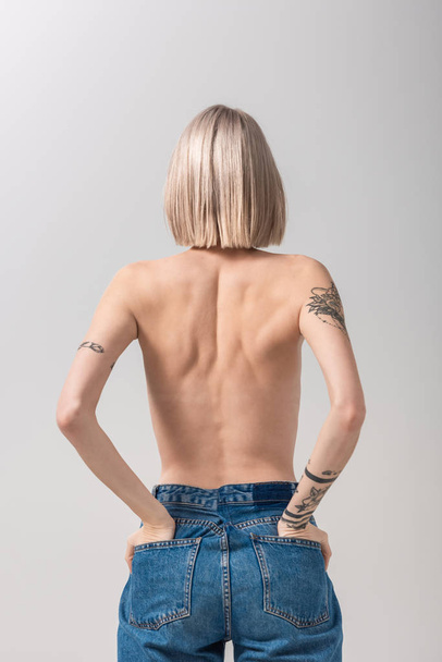hátsó kilátás szexi fiatal topless tetovált nő pózol a kezét zsebében elszigetelt szürke - Fotó, kép