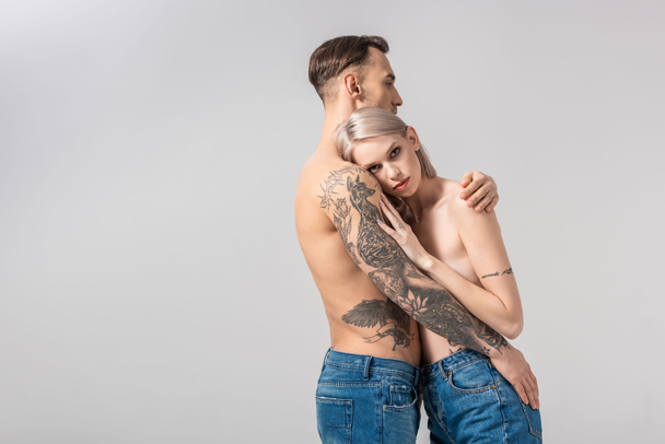 вид сбоку на молодую обнаженную татуированную пару, обнимающую изолированную на сером
 - Фото, изображение