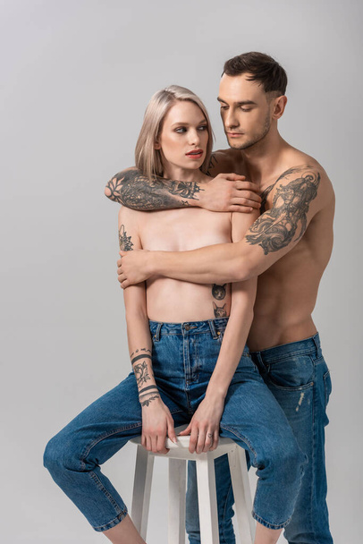 young shirtless tattooed couple in jeans hugging on chair isolated on grey - Foto, imagen