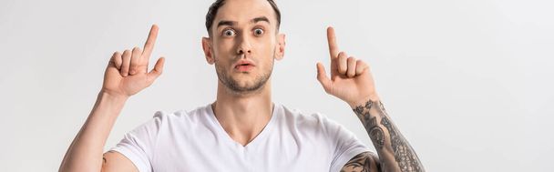 shocked handsome young tattooed man pointing with fingers up isolated on white, panoramic shot - Photo, Image