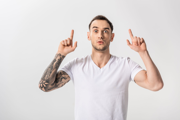 sorprendido guapo joven tatuado hombre señalando con los dedos hacia arriba aislado en blanco
 - Foto, Imagen