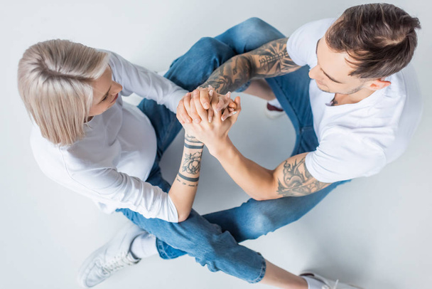 overhead view of young tattooed pregnant woman sitting with husband on floor and holding hands on grey - Photo, Image