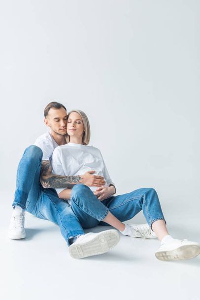 young tattooed pregnant woman with closed eyes sitting with husband on floor on grey - Foto, immagini