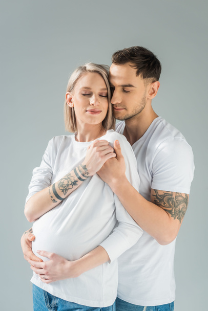 молодая татуированная беременная женщина рядом с парнем с закрытыми глазами, изолированными на серой
 - Фото, изображение