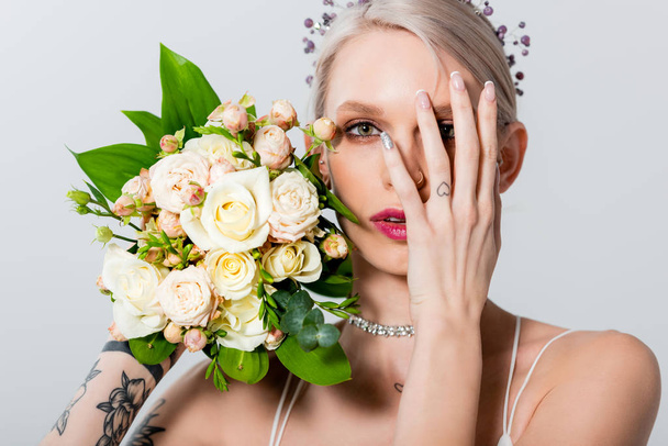 portrait of beautiful tattooed bride posing with floral bouquet and hand on face isolated on grey - Foto, Bild