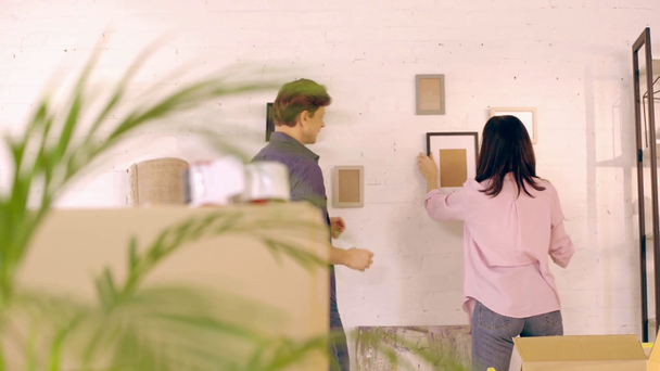 Couple hanging empty photo frames on wall near packages - Video, Çekim