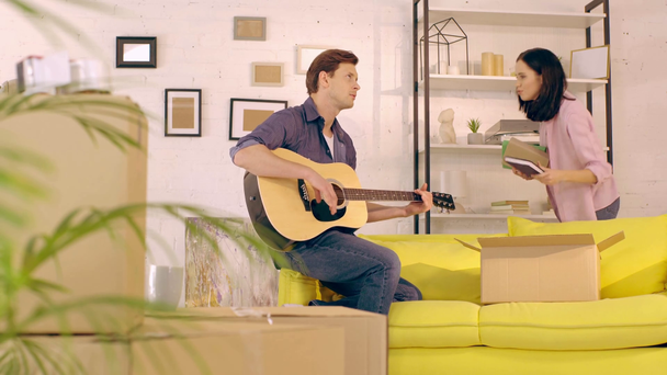 Mädchen packt Paket in der Nähe ihres Freundes aus und spielt Gitarre im neuen Zuhause - Filmmaterial, Video