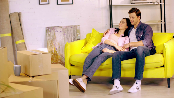 Χαμογελαστό ζευγάρι που μιλάει στον καναπέ κοντά σε πακέτα στο νέο σπίτι - Πλάνα, βίντεο