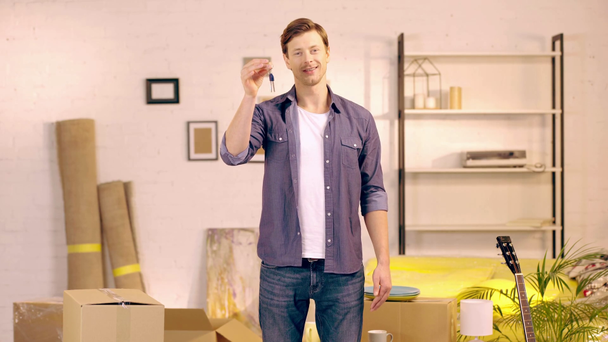 Uśmiechnięty mężczyzna pokazuje klucze w pobliżu pakietów w nowym domu - Materiał filmowy, wideo