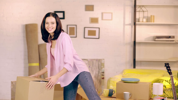 Χαμογελώντας κορίτσι βάζοντας πακέτο και χαμογελώντας στην κάμερα στο νέο σπίτι - Πλάνα, βίντεο