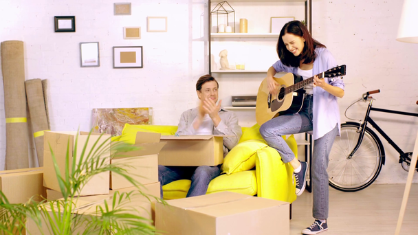 Dziewczyna gra na gitarze w pobliżu chłopaka z pudełka na kanapie w nowym domu - Materiał filmowy, wideo