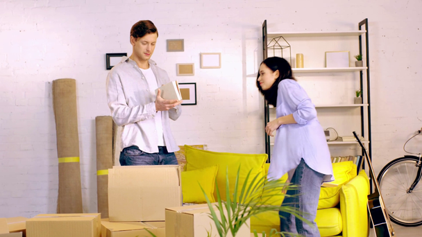 Νεαρό ζευγάρι συσκευάζει βιβλία σε κουτιά στο σαλόνι - Πλάνα, βίντεο