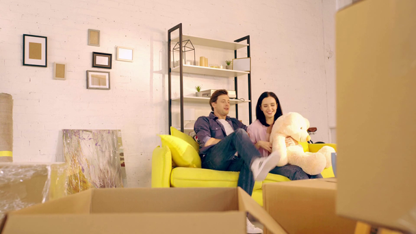 Мужчина берет плюшевого мишку из упаковки и дает девушке на диване
  - Кадры, видео