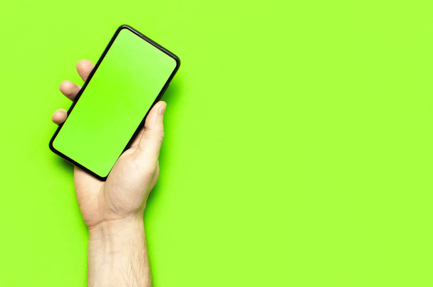 Las manos masculinas sostienen un teléfono inteligente negro moderno con pantalla en blanco verde sobre fondo verde neón plano vista superior. Tecnología moderna, teléfono, gadget en manos, pantalla táctil, plantilla para su diseño. Burla.
 - Foto, Imagen