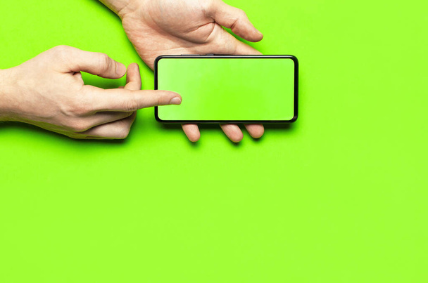 Чоловічі руки тримають сучасний чорний смартфон із зеленим порожнім екраном на неоновому зеленому фоні з плоским видом зверху. Сучасні технології, телефон, гаджет в руках, сенсорний екран, шаблон вашого дизайну. Мокап
 - Фото, зображення