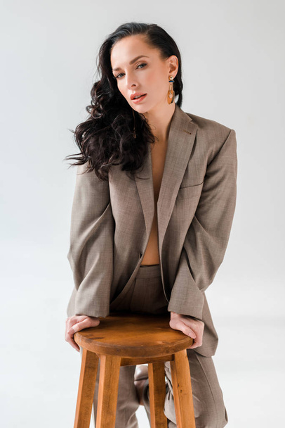 stylish woman in suit on stool looking at camera on grey background  - Foto, Imagem