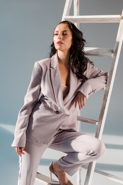 stylish woman in suit near ladder looking away on grey background  - Zdjęcie, obraz