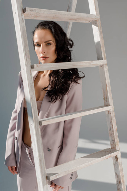 sexy and stylish woman in suit near ladder looking at camera on grey background  - Foto, Bild