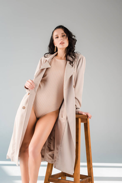 attractive woman in bodysuit and coat standing near wooden stool on grey background  - Fotó, kép