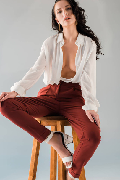 attractive and sexy woman in shirt and trousers sitting on stool isolated on grey - Foto, Bild
