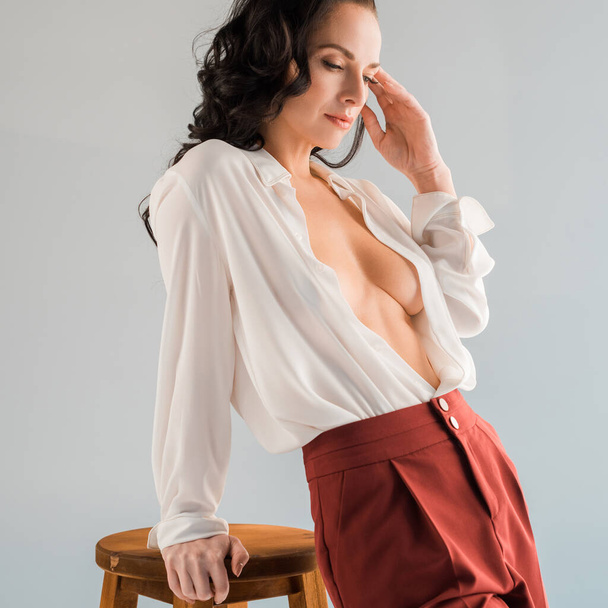 привлекательная и сексуальная женщина в рубашке и брюках, стоящих возле стула, изолированного на сером
  - Фото, изображение