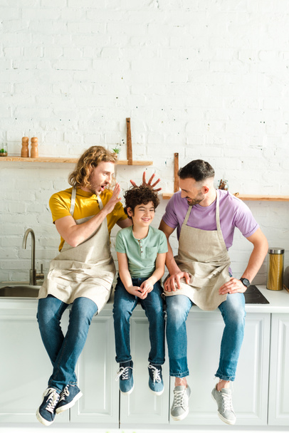 happy homosexual parents sitting with mixed race son in kitchen - Fotoğraf, Görsel