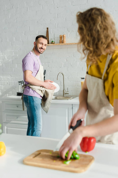 επιλεκτική εστίαση του χαρούμενου ομοφυλόφιλου άνδρα κοιτάζοντας σγουρά σύντροφο, ενώ πλύσιμο πλάκα  - Φωτογραφία, εικόνα