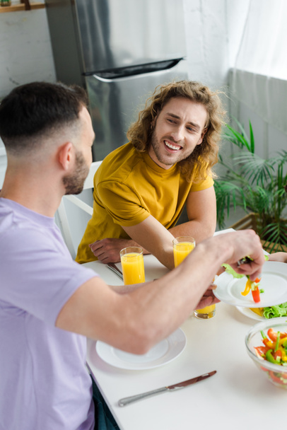 homosexual and bearded man putting salad on plate of happy husband  - Fotografie, Obrázek