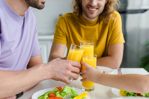 cropped view of happy homosexual men and son clinking glasses with orange juice  - Photo, Image