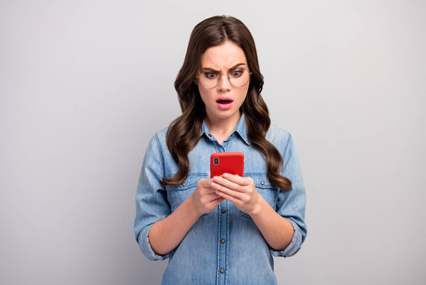 Photo of beautiful freelancer shocked lady hold browsing telephone check followers open mouth bad news wear specs casual jeans denim shirt isolated grey color background - Foto, afbeelding