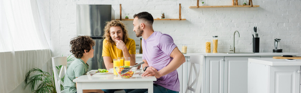 plano panorámico de los padres homosexuales y lindo hijo de raza mixta almorzando en casa
 - Foto, Imagen