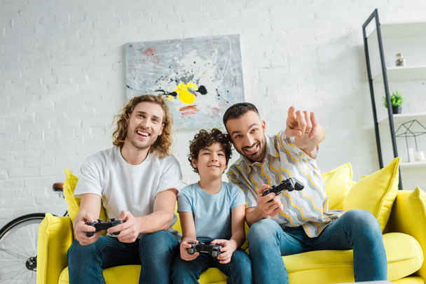 KYIV, UKRAINE - JANUARY 28, 2020: cheerful homosexual couple playing video game with mixed race son  - Фото, изображение