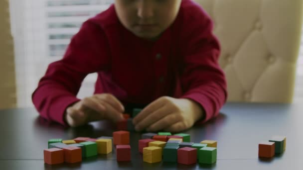 small boy in a red sweater is playing with the colorful wooden blocks on a table in front of a window.Closeup video. - Кадри, відео