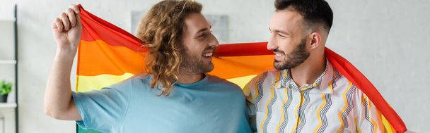 панорамний знімок щасливих гомосексуальних чоловіків, що тримають прапор ЛГБТ і дивляться один на одного
  - Фото, зображення