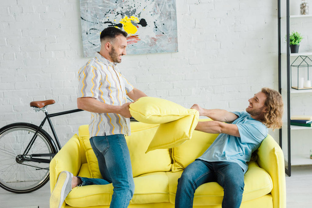 vista lateral de homens homossexuais felizes travesseiro lutando na sala de estar
  - Foto, Imagem